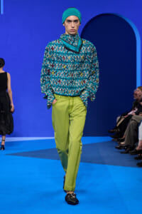 Model walks the runway in a turquoise beanie, teal patterned top, and lime-green trousers on a blue set.
