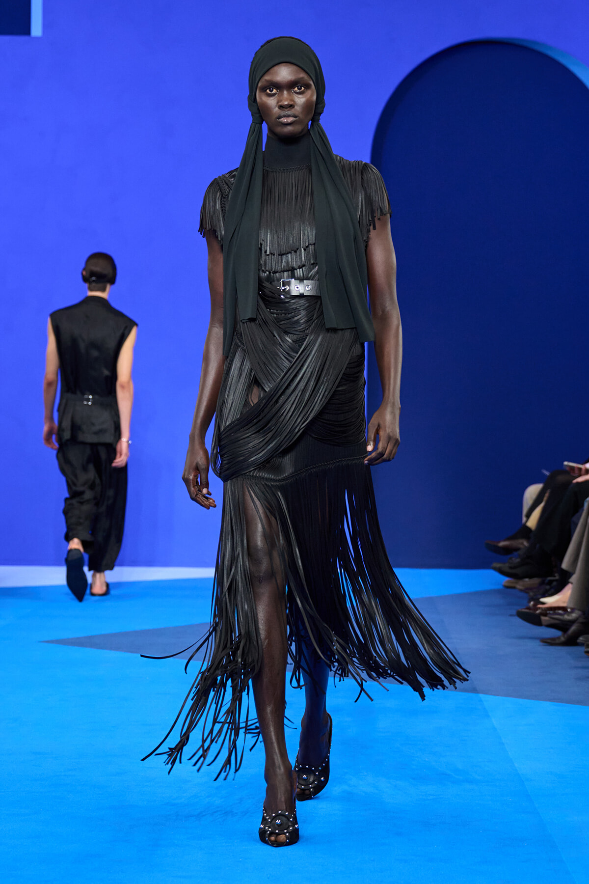 Model in a black layered fringe dress and headscarf walks a blue runway at a fashion show.