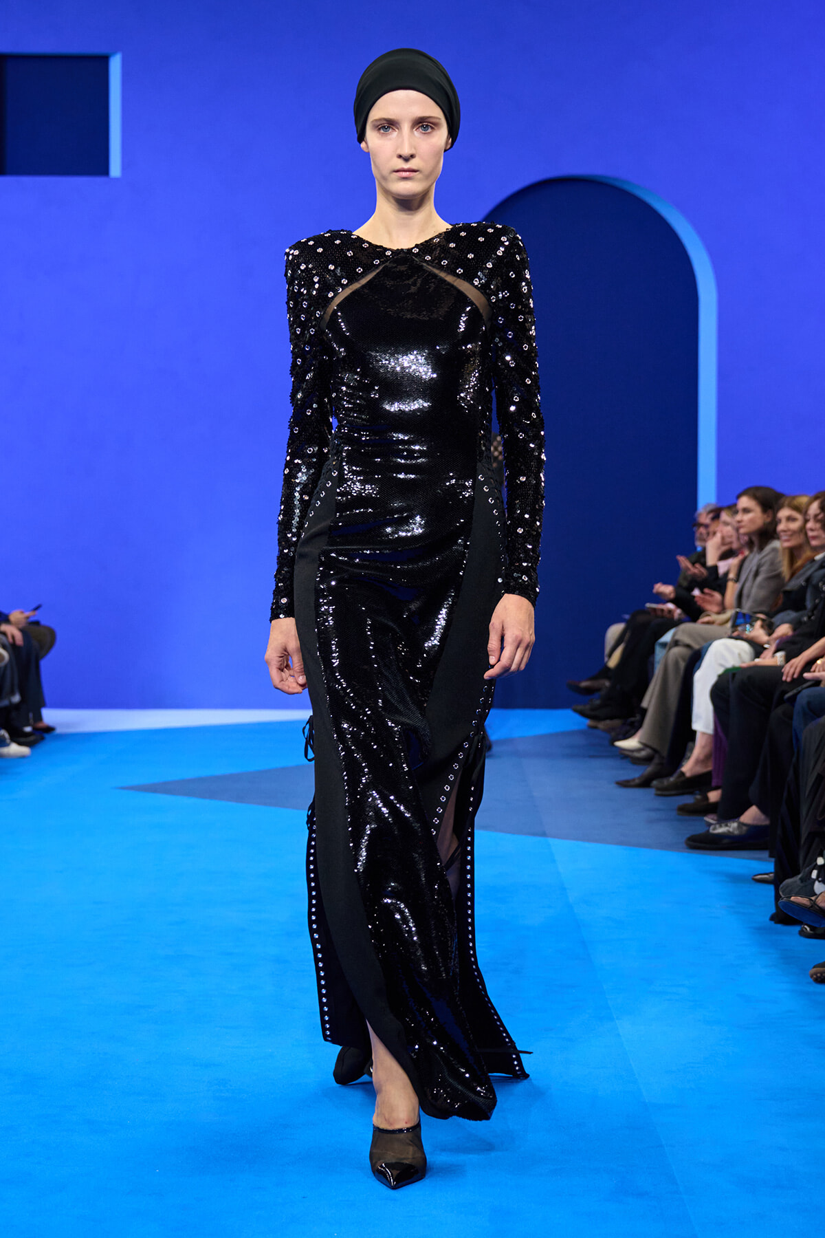 Model walks a black sequined gown with sheer panels on a bright blue runway, wearing a black head wrap.