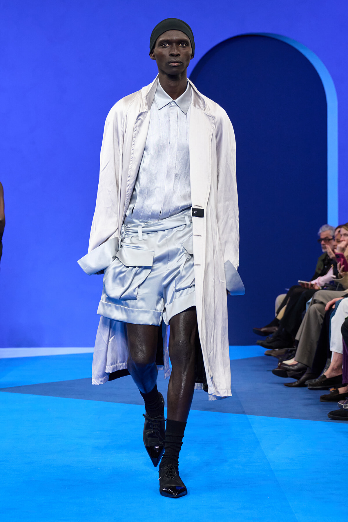 Male model walks a blue runway in a pale satin shirt and shorts set, paired with a long beige coat and black boots, during a fashion show.