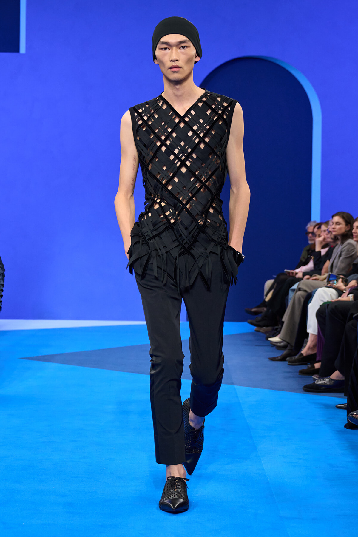 Male runway model in a black geometric cutout top and beanie walking the blue catwalk with seated audience watching the show.