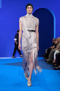 Model on a blue runway wearing a silver fringe gown with a high neck, black belt, and draped fringe detailing; silver platform heels and a black headscarf.