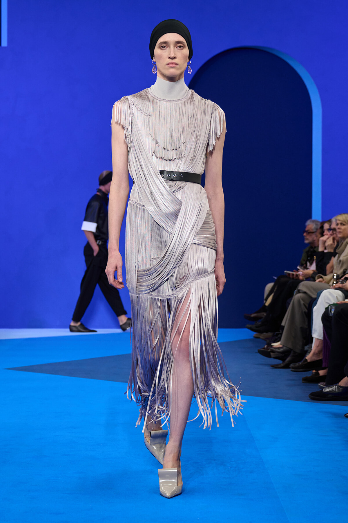 Model on a blue runway wearing a silver fringe gown with a high neck, black belt, and draped fringe detailing; silver platform heels and a black headscarf.
