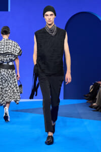 Male model on a blue runway in an all-black ensemble: draped sleeveless top, slim trousers, black head wrap, and layered silver neckwear.