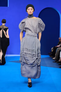 Model walks a pale blue, gathered-ruffle dress on a bright blue runway, wearing a black head wrap and dark footwear.