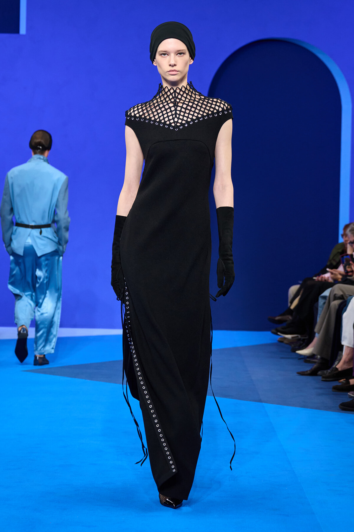 Model in a black floor-length dress with a perforated lattice neckline, black head wrap, and long gloves on a blue runway surface.