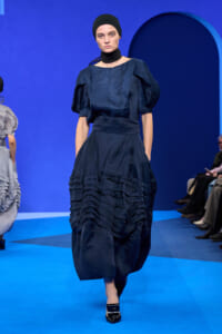 Model walking a blue runway in a dark blue, voluminous, ruffled gown with a matching sash and black head wrap; poised expression.