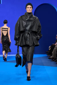 Model walking a fashion runway in a textured black leather oversized belted jacket, matching skirt, and black head wrap, carrying a black handbag on a blue-lit catwalk.