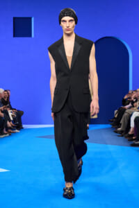 Model walks the fashion runway in a sleeveless black blazer over a tan top, wide black pants, and a jeweled black cap; blue set background.