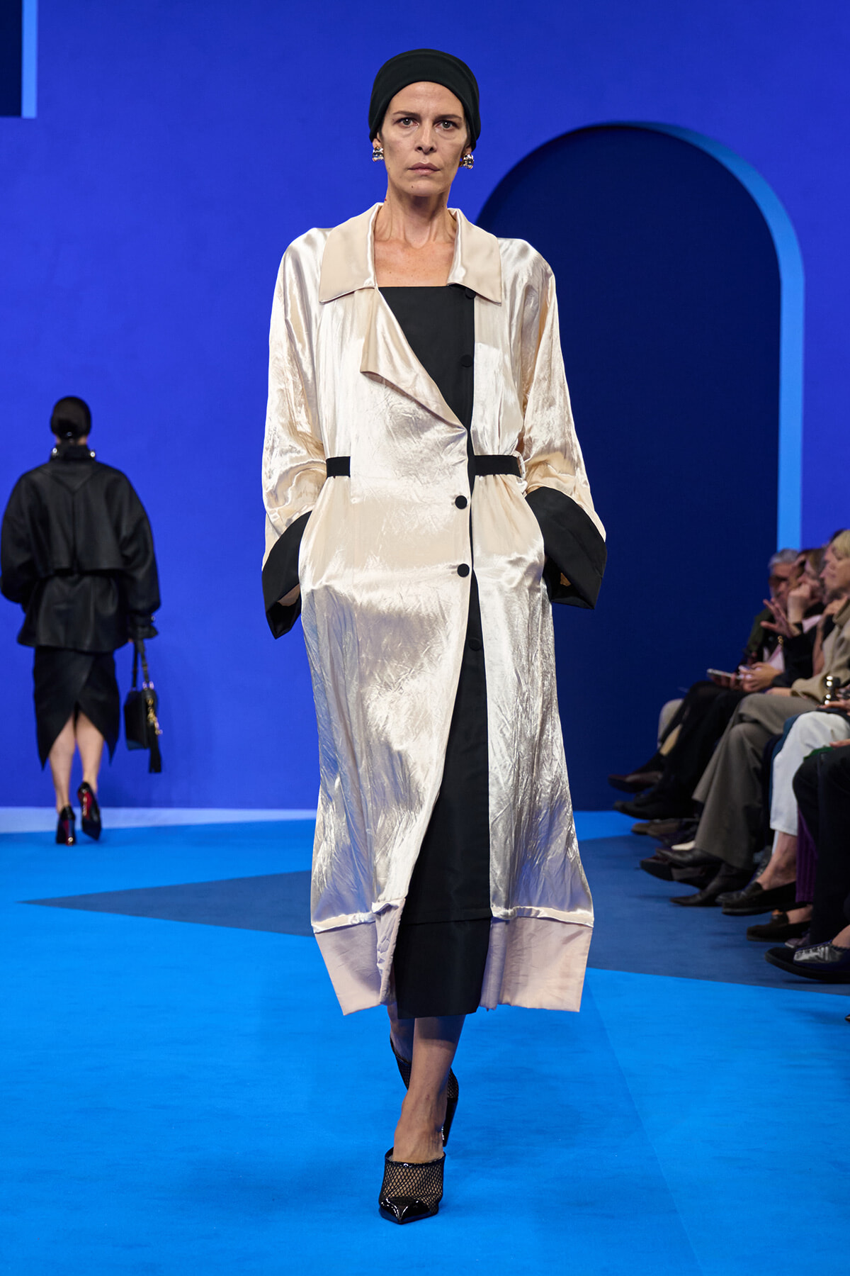 Female model walks the runway in a shiny champagne trench coat over a black dress, with a black headscarf and pointed heels on a blue catwalk.