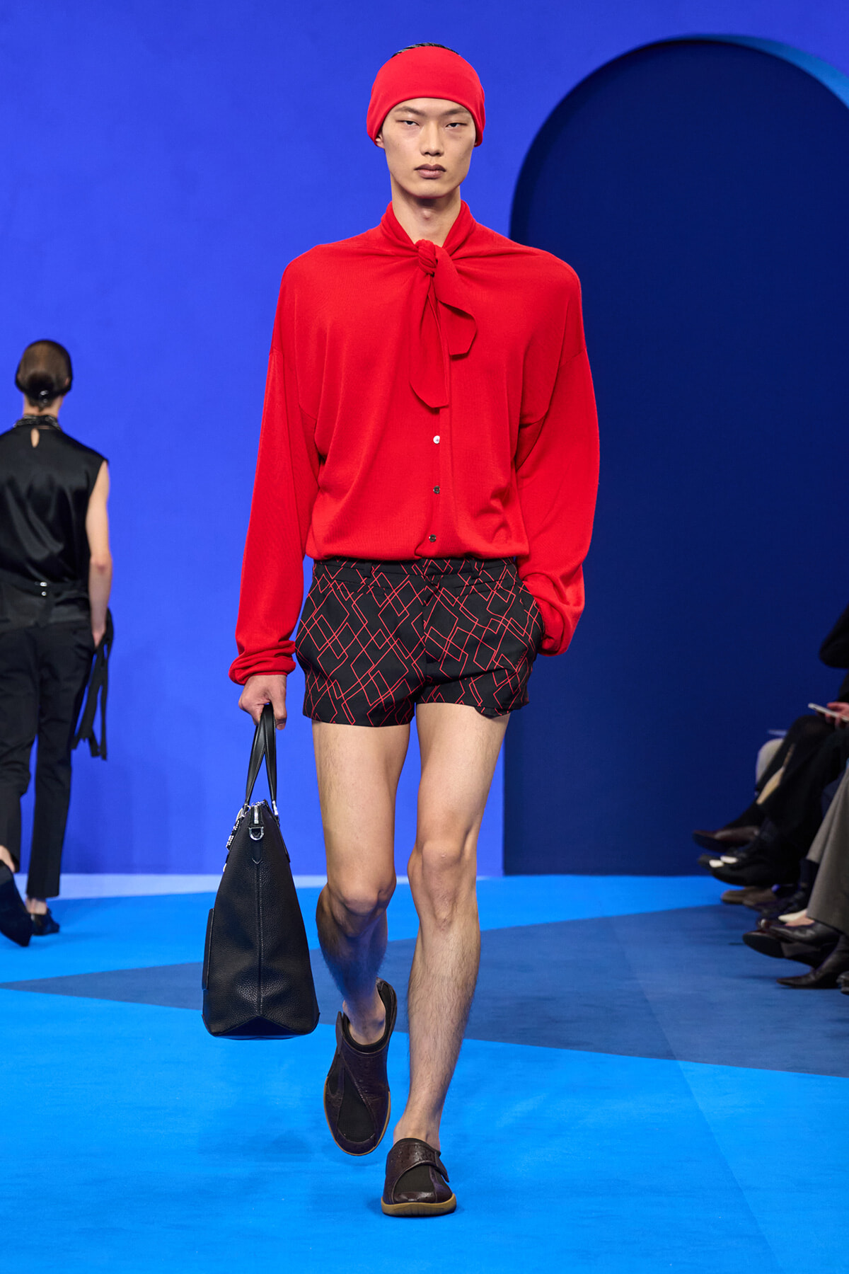 Male runway model in a red blouse and headscarf, black geometric shorts, carrying a black handbag on a blue catwalk.