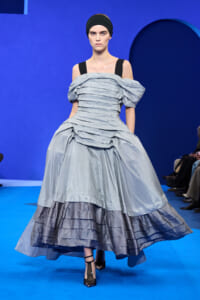 Model on a blue runway wearing an off-shoulder light-blue ballgown with layered, crinkled fabric and a black headband, black ankle-strap heels