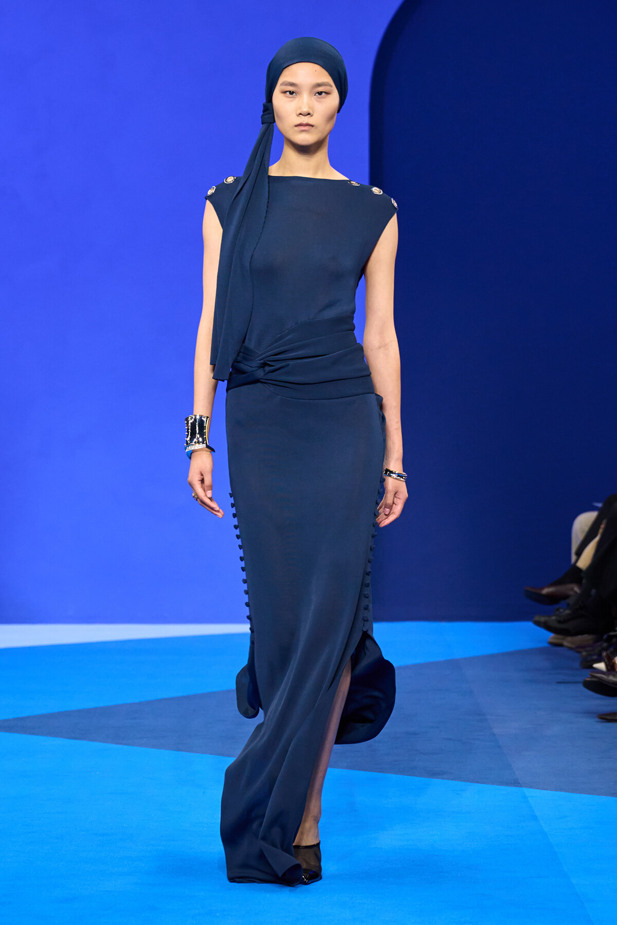Model walking a dark blue sleeveless gown with a twisted waist, matching headscarf, and silver bracelets on a blue runway.