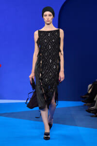 Model walking a runway in a sleeveless black sequined dress with diamond lattice pattern and side ribbon fringe, wearing a black headwrap against a bright blue backdrop.