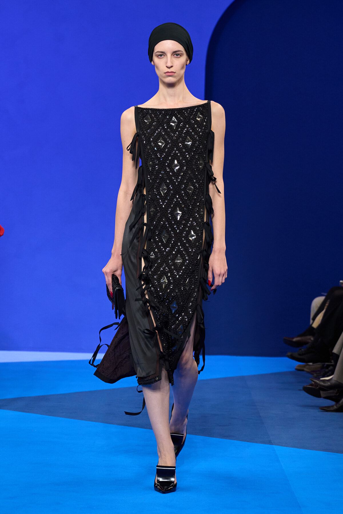 Model walking a runway in a sleeveless black sequined dress with diamond lattice pattern and side ribbon fringe, wearing a black headwrap against a bright blue backdrop.