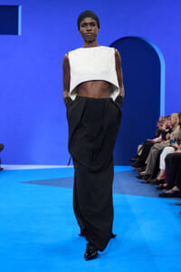 Model walks a fashion show wearing a white sleeveless cropped top and black wide-leg pants on a blue runway with seated audience in the background, hands in pockets.