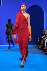 Model in a red one-shoulder dress with a matching headscarf walking a fashion runway against a blue backdrop, holding a red clutch.