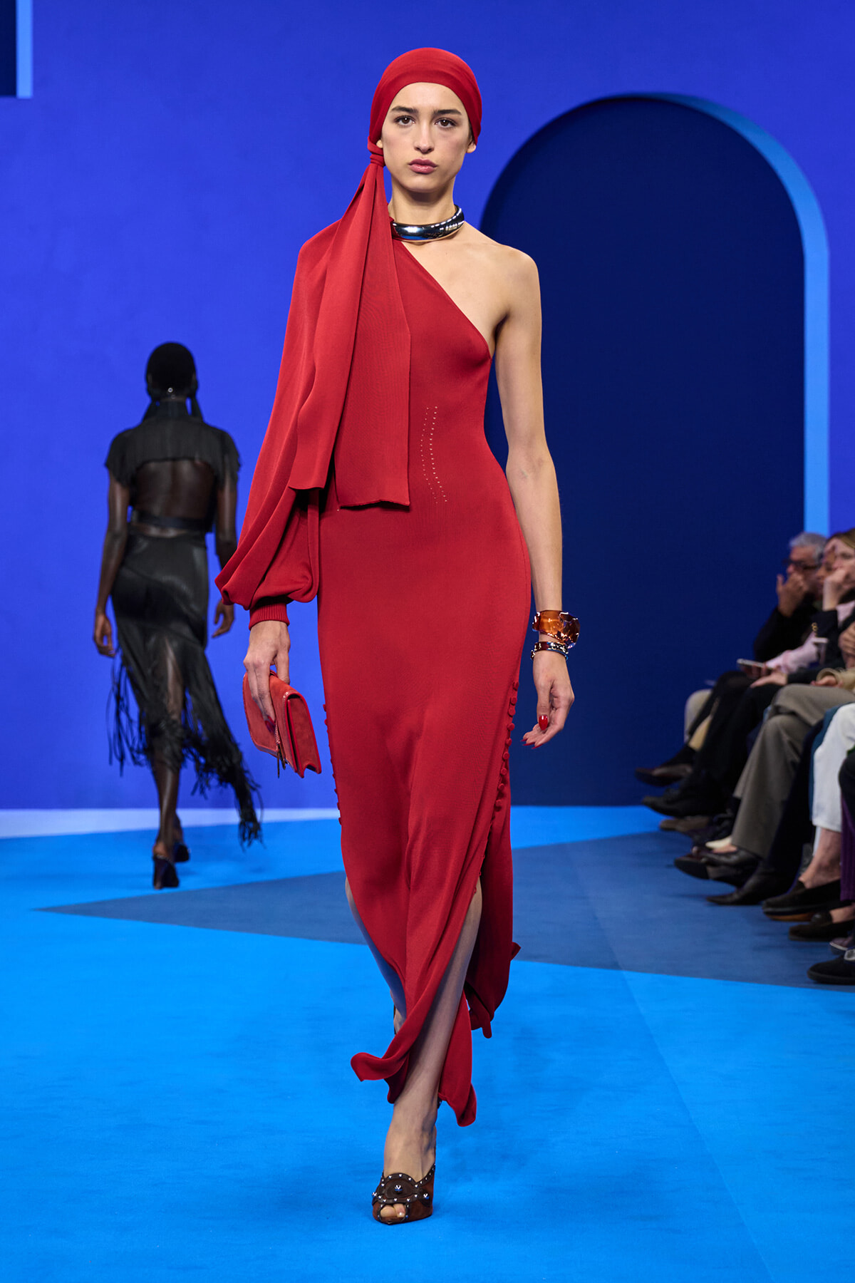 Model in a red one-shoulder dress with a matching headscarf walking a fashion runway against a blue backdrop, holding a red clutch.