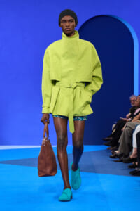 Model walks a bright lime-green oversized trench with a tied waist on a blue runway; turquoise loafers and a brown handbag complete the look.
