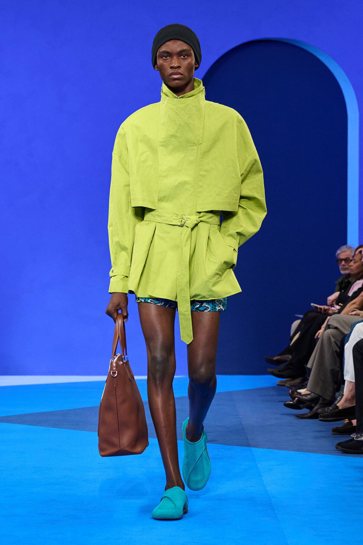 Model walks a bright lime-green oversized trench with a tied waist on a blue runway; turquoise loafers and a brown handbag complete the look.