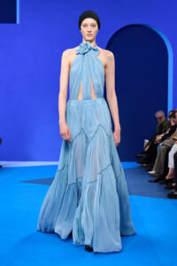 Model walks the runway in a light blue halter dress with draped pleats and cutouts at the bodice, wearing a black head wrap.