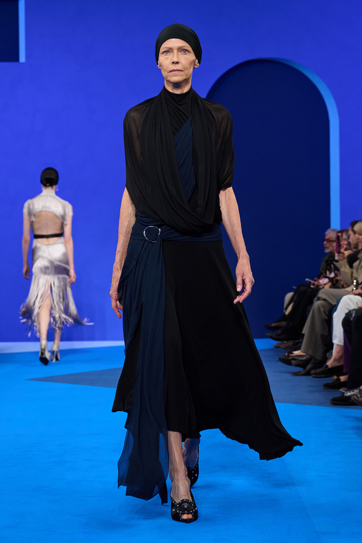 Runway model in a black hijab and draped black and navy outfit walking a blue catwalk with spectators on the side.