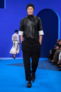 Male model walking a fashion runway in a black leather top, black utility pants, and a black beanie against a blue set.
