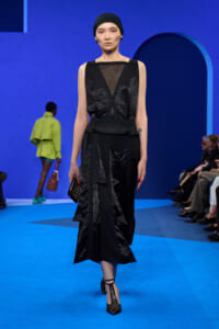 Model walks a runway wearing a black sleeveless dress with mesh neckline, crisscross belt, head wrap, and black heels against a bright blue backdrop.