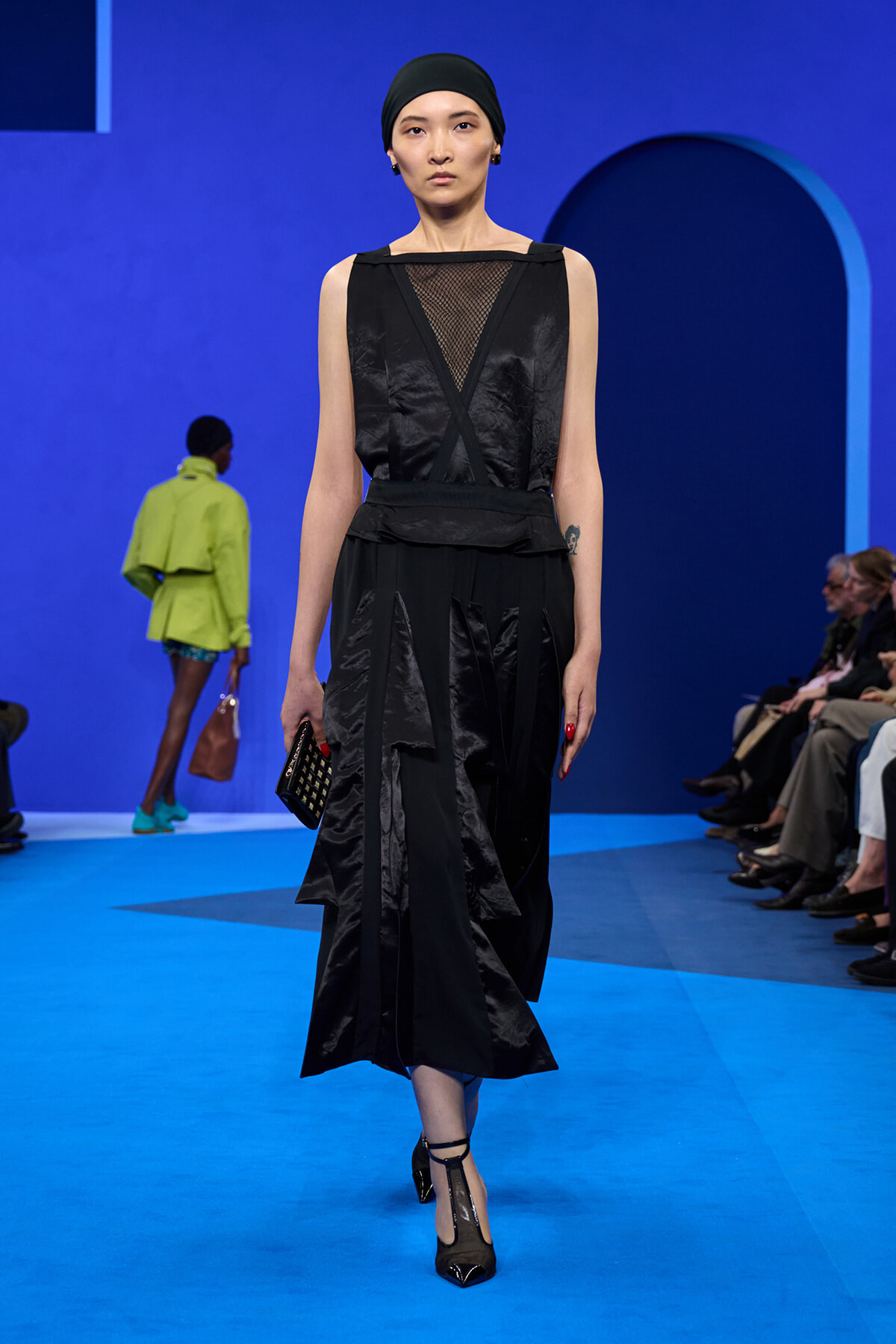 Model walks a runway wearing a black sleeveless dress with mesh neckline, crisscross belt, head wrap, and black heels against a bright blue backdrop.