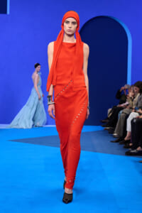 Model walks the runway in a vivid orange sleeveless dress with matching head scarf against a blue set, audience watching on the right.