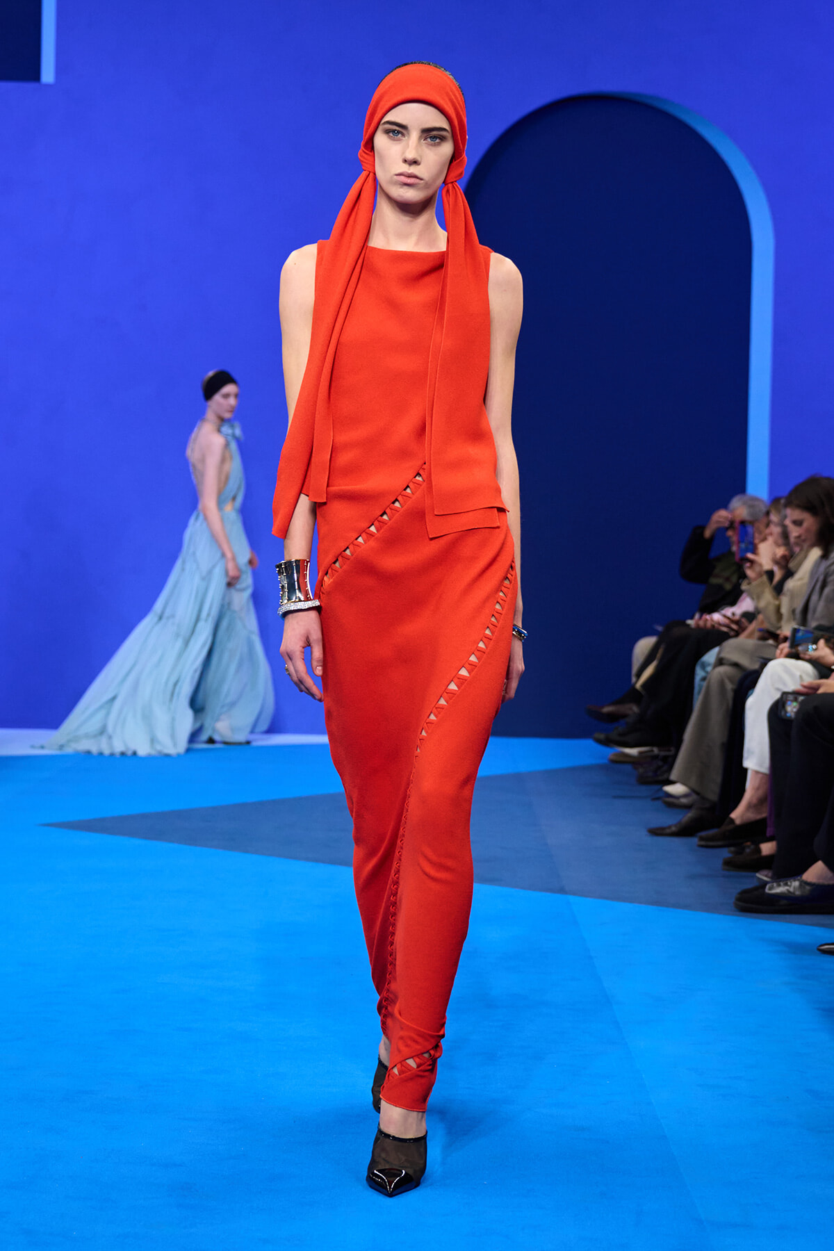 Model walks the runway in a vivid orange sleeveless dress with matching head scarf against a blue set, audience watching on the right.