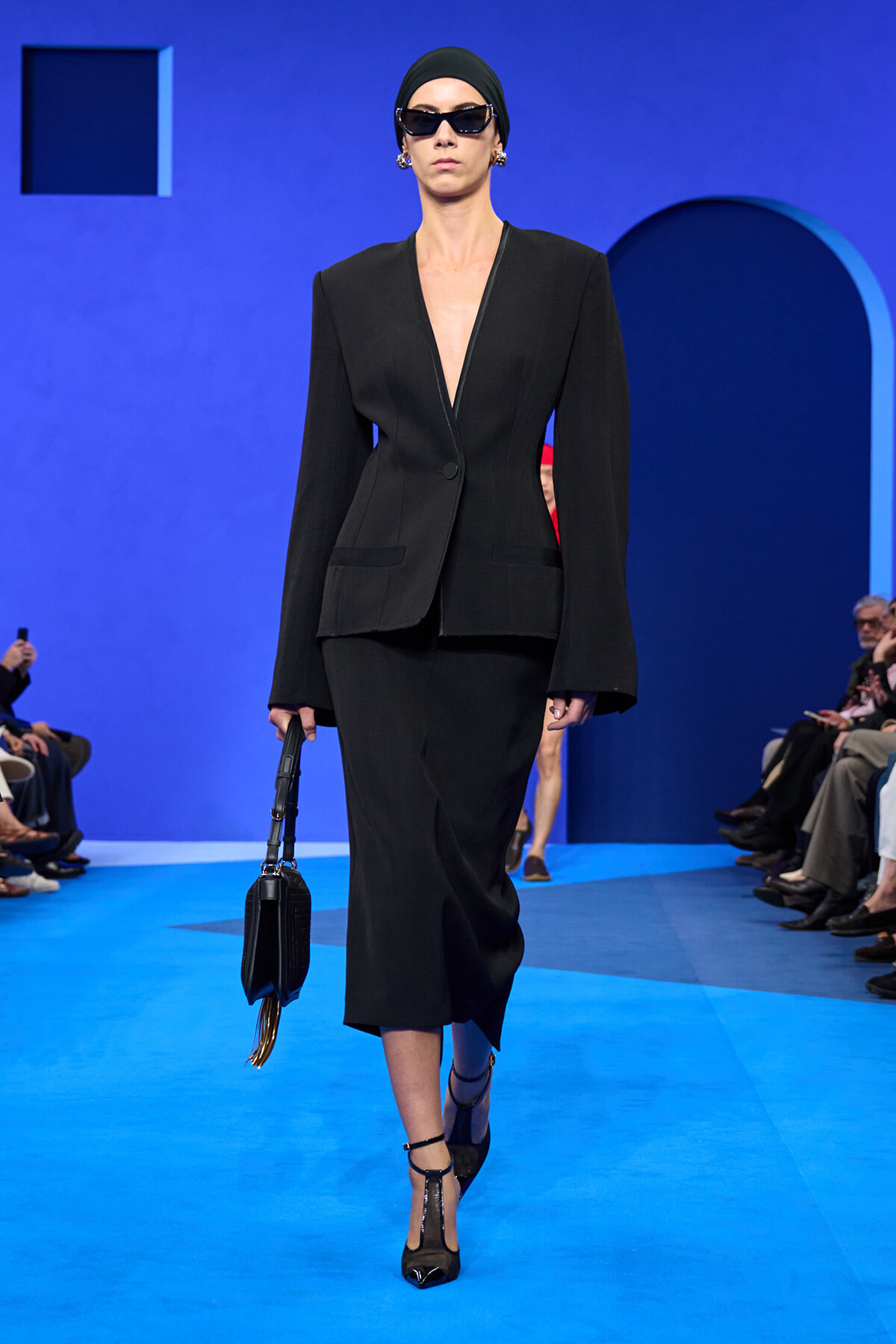 Model walking the runway in a black tailored blazer and matching skirt, wearing a black headscarf and oversized sunglasses, carrying a black handbag on a blue set.