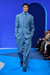 Male model in a light blue suit with a matching scarf, black beanie, walking a blue runway with audience on the side.
