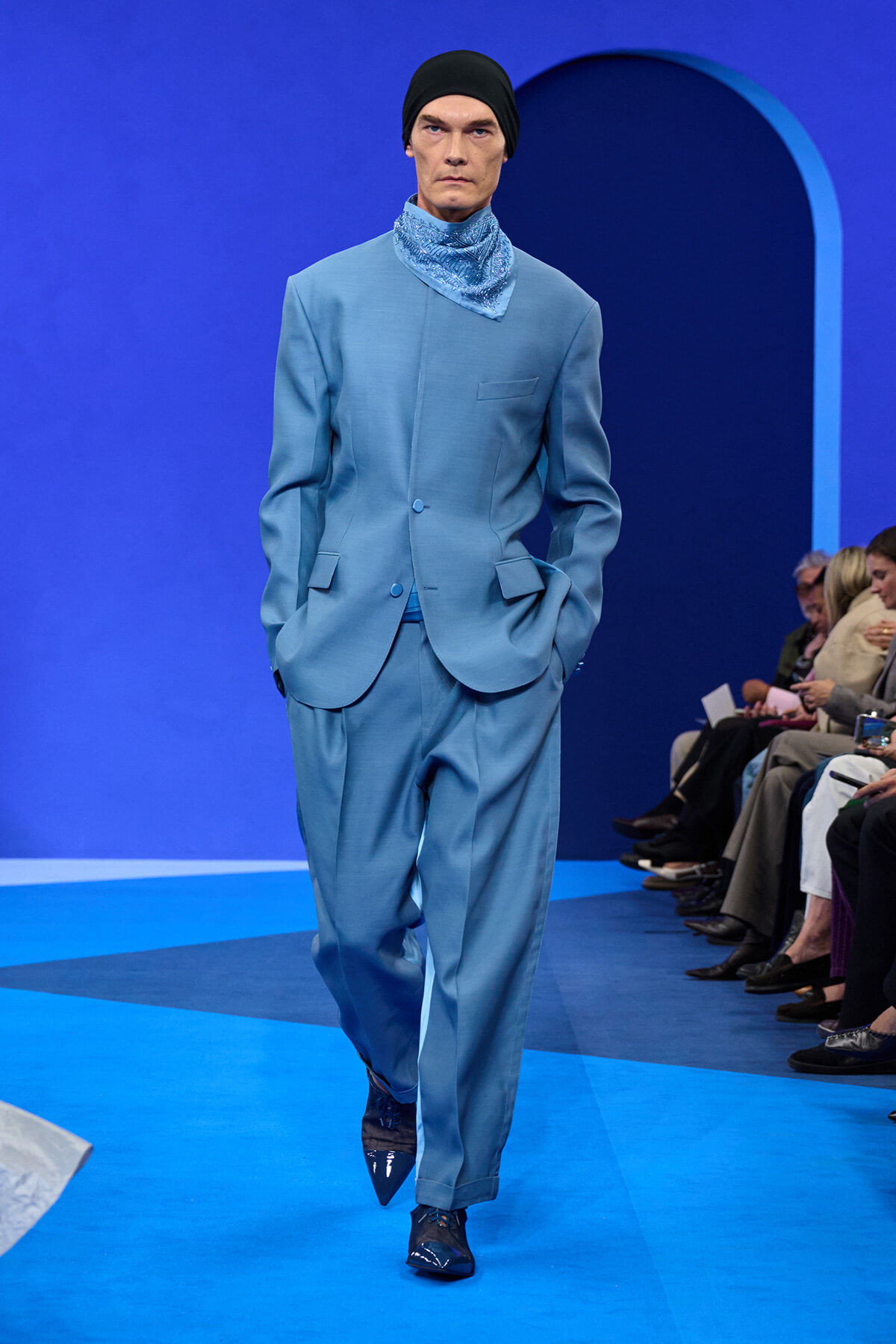 Male model in a light blue suit with a matching scarf, black beanie, walking a blue runway with audience on the side.