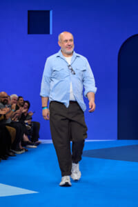 Man walking a blue runway wearing a light blue shirt over a white tee, dark pants, white sneakers, with glasses hanging from his chest; audience applauds at left.