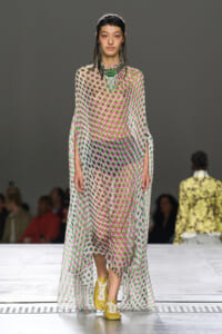 Model on a runway wearing a sheer pink‑green polka‑dot maxi dress with cape sleeves, a bold green necklace, and yellow sneakers.