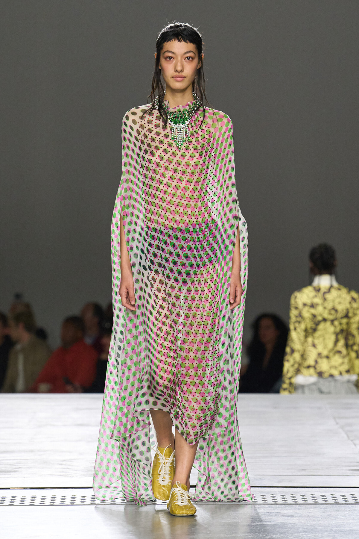 Model on a runway wearing a sheer pink‑green polka‑dot maxi dress with cape sleeves, a bold green necklace, and yellow sneakers.