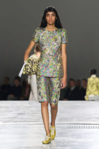 Female model walks a runway in a metallic multicolor brocade short-sleeve top and matching knee-length shorts, carrying a patterned handbag under her arm.