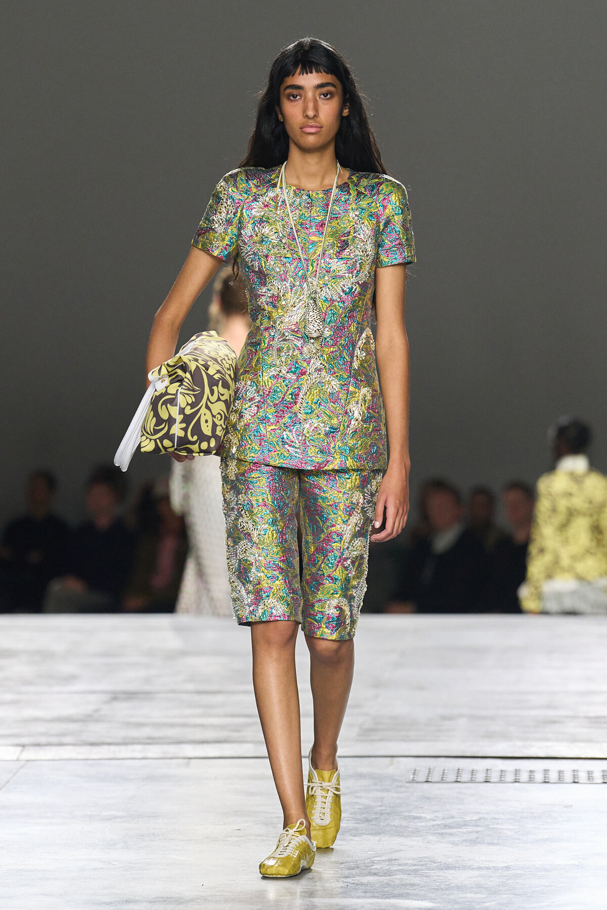 Female model walks a runway in a metallic multicolor brocade short-sleeve top and matching knee-length shorts, carrying a patterned handbag under her arm.