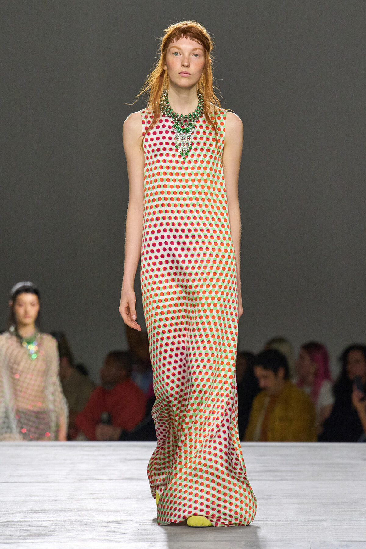 Model walking the runway in a sleeveless white jumpsuit covered in red-green polka dots, paired with a chunky green necklace and yellow shoes.