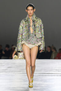Model walks the runway in a metallic, floral jacket over a neon pink and green ruffled blouse with beige shorts and yellow sneakers on a grey catwalk background.