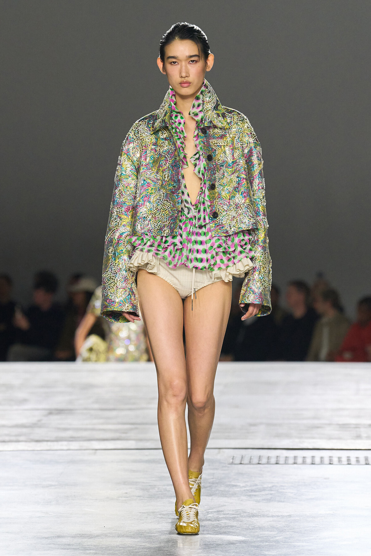 Model walks the runway in a metallic, floral jacket over a neon pink and green ruffled blouse with beige shorts and yellow sneakers on a grey catwalk background.