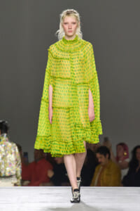 Model on a runway wearing a bright lime-green sheer polka-dot dress with layered ruffles and black lace-up ankle boots.