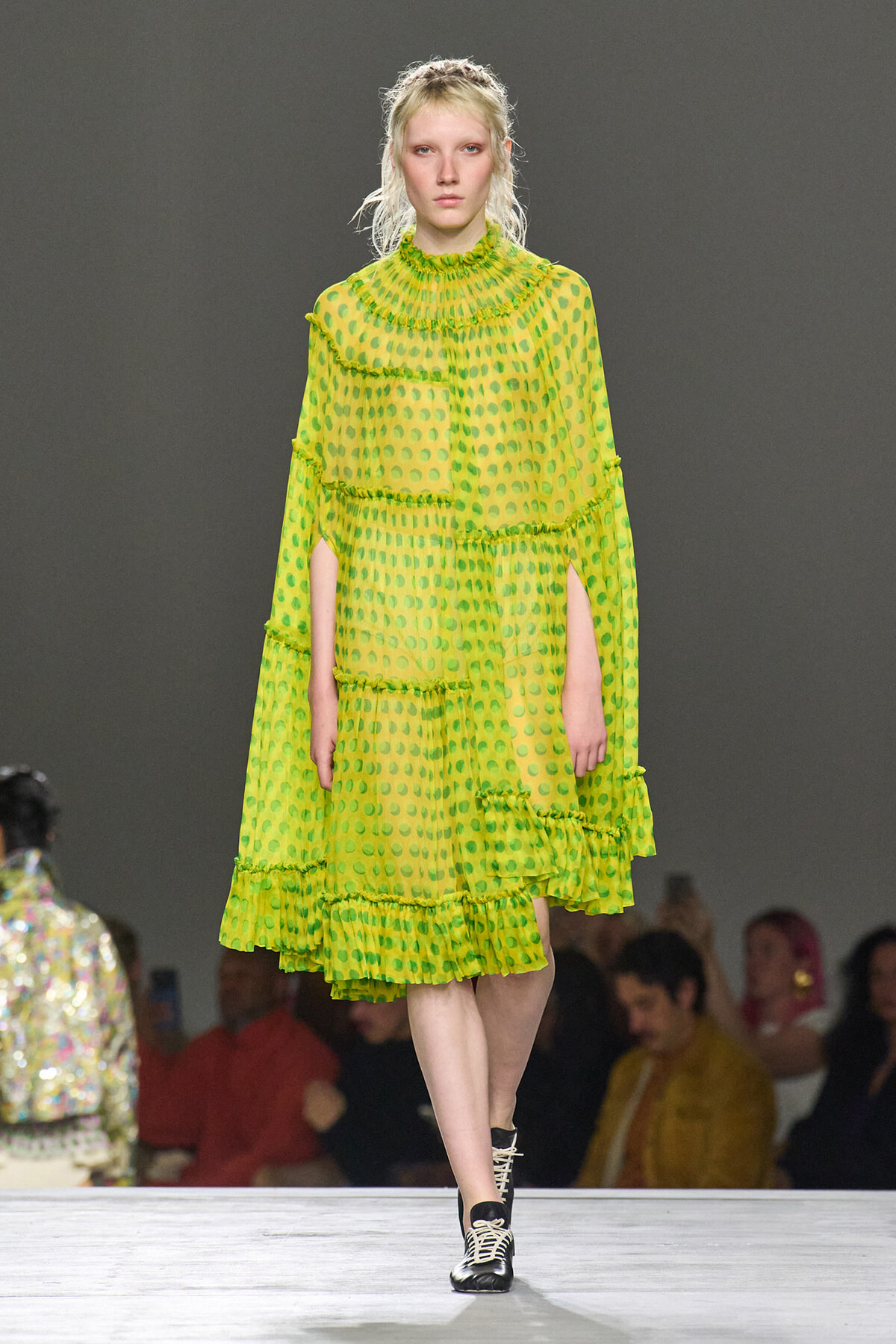 Model on a runway wearing a bright lime-green sheer polka-dot dress with layered ruffles and black lace-up ankle boots.
