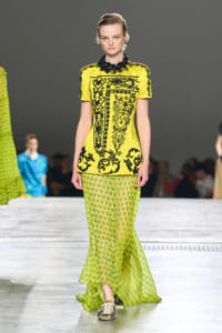 Model walks the runway in a bright yellow embroidered top paired with a lime green polka-dot skirt; fashion show setting in background.