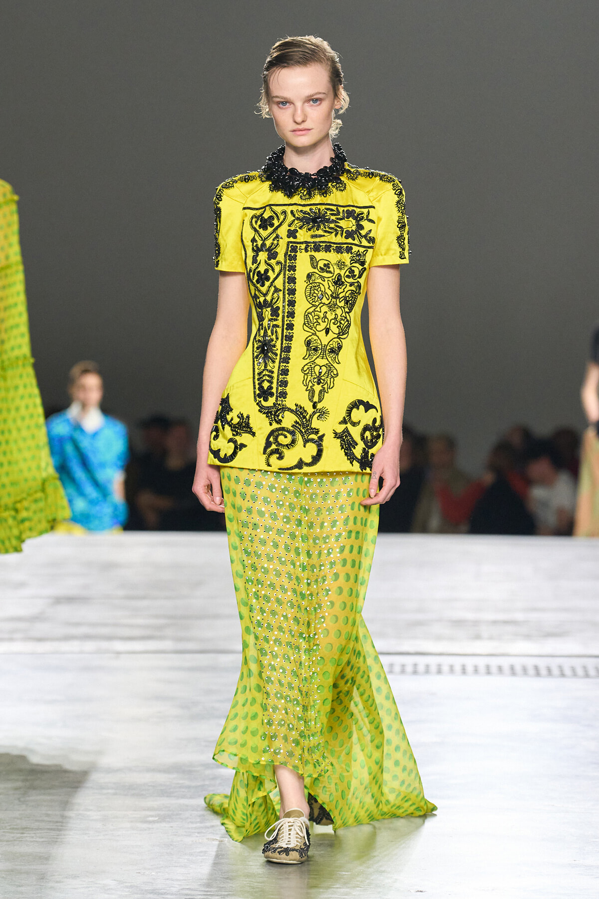 Model walks the runway in a bright yellow embroidered top paired with a lime green polka-dot skirt; fashion show setting in background.
