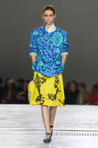 Female model walks a runway wearing a bright blue floral blazer and a yellow embroidered skirt.