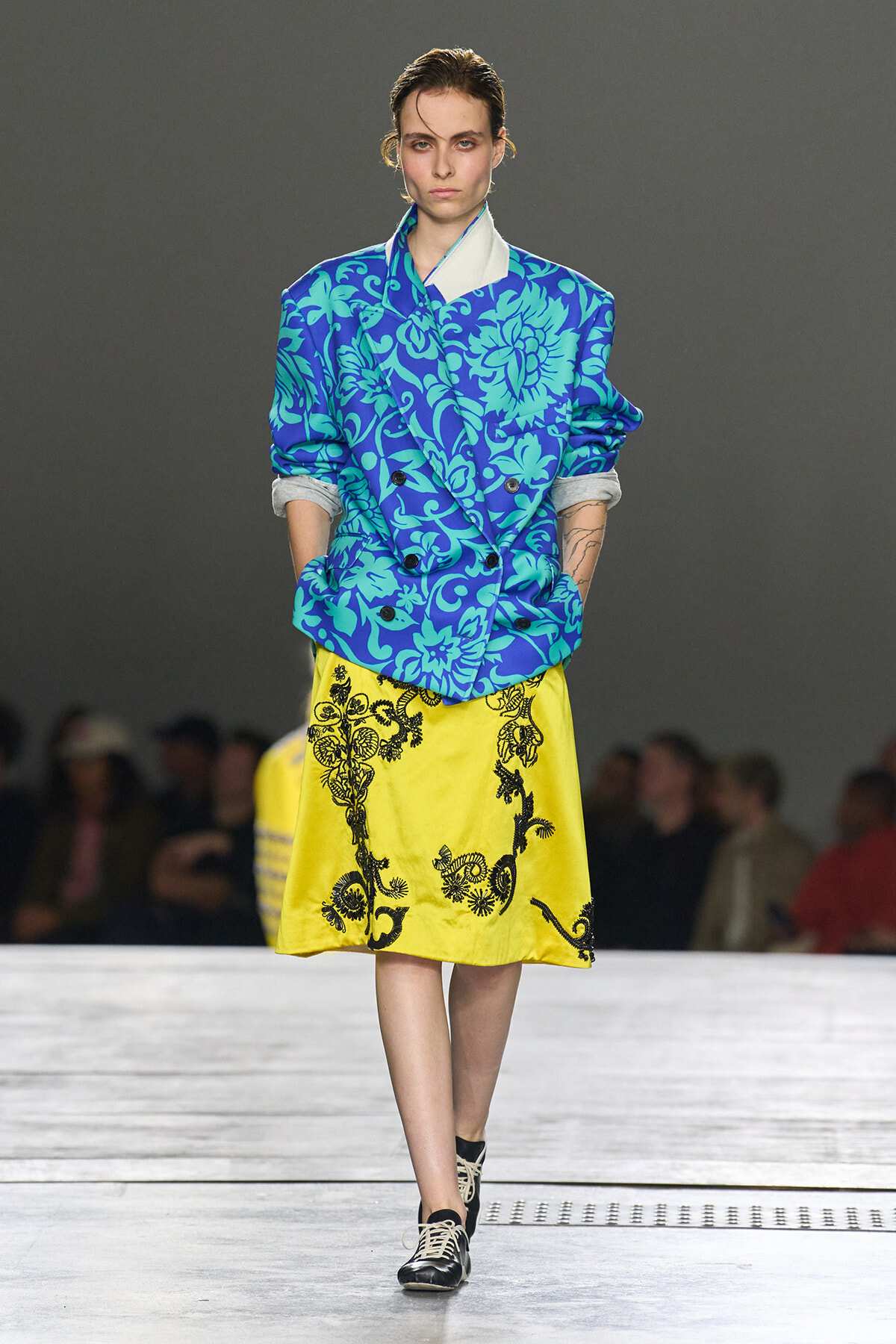 Female model walks a runway wearing a bright blue floral blazer and a yellow embroidered skirt.