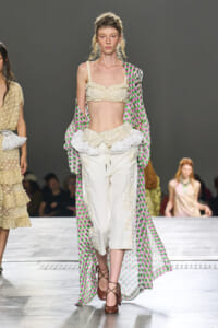 Runway model in a cream ruffled crop top and embroidered wide-leg pants with a sheer pink–green polka-dot cape draped over her shoulders.
