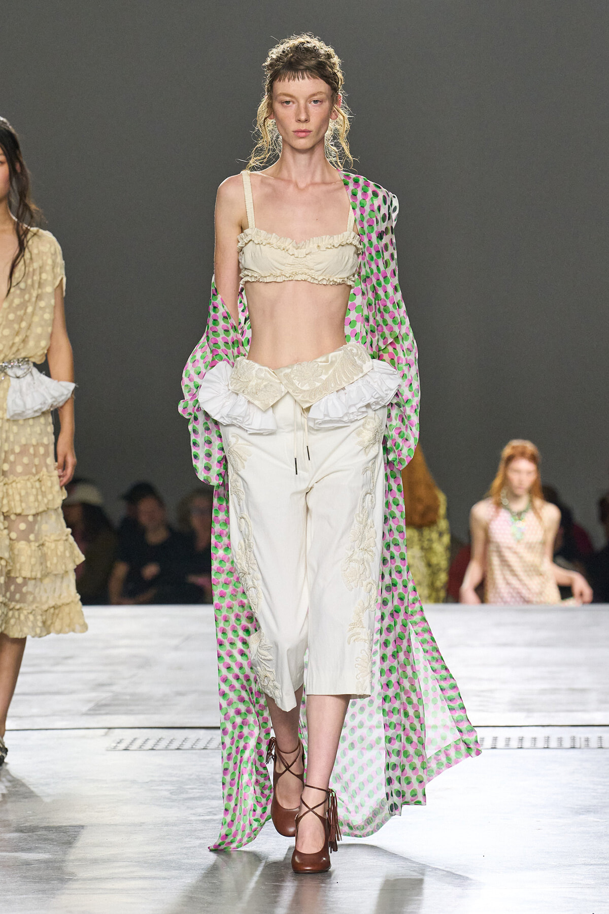 Runway model in a cream ruffled crop top and embroidered wide-leg pants with a sheer pink–green polka-dot cape draped over her shoulders.